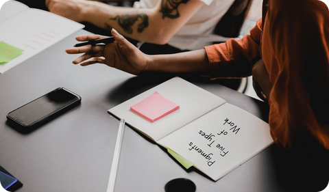 Two colleagues in a collaborative discussion at a meeting table. An open notebook displays "Pigment's Five Types of Work" with pink sticky notes, alongside a smartphone. One person wears an orange shirt while the other, in white, gestures with tattooed ar
