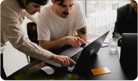 Three colleagues collaborate around a table covered with papers, magazines, sticky notes, and documents. A woman with dark bangs in a black sweater sits at center, resting her chin on her hand while reviewing materials. Two others lean in from either side