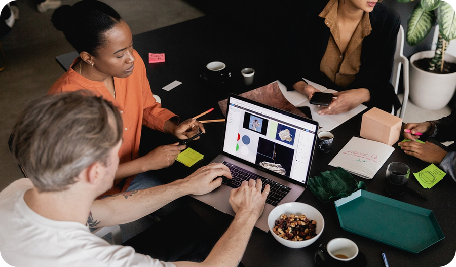 Alt text: Overhead view of a team collaborating around a dark table. One person types on a laptop showing a design presentation while a colleague in an orange shirt points with a pencil. Coffee cups, sticky notes, notebooks, and a bowl of snacks are scatt