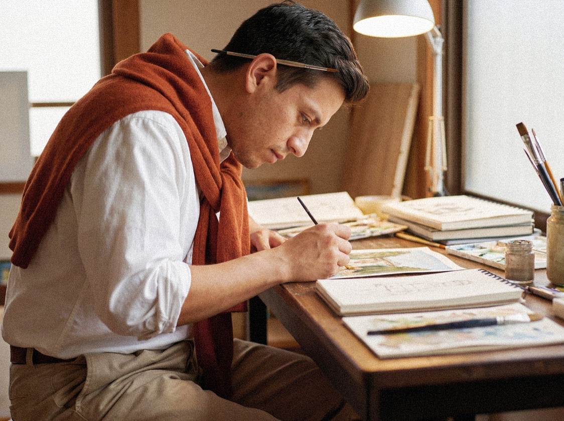 Person drawing with brushes in sketchbooks at a warmly lit wooden desk.