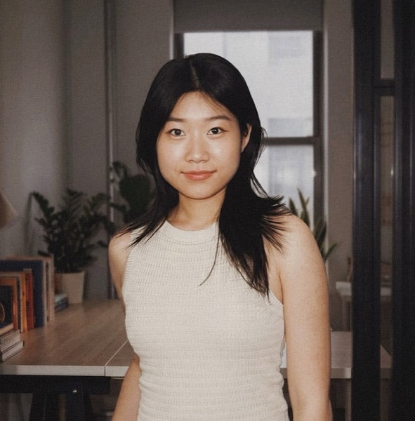 Ada Zhou, long dark hair in a cream top, standing in a plant-filled office by a window