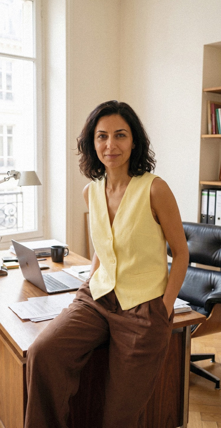 Woman in a yellow vest sitting on a desk in a sunlit office, calm and confident