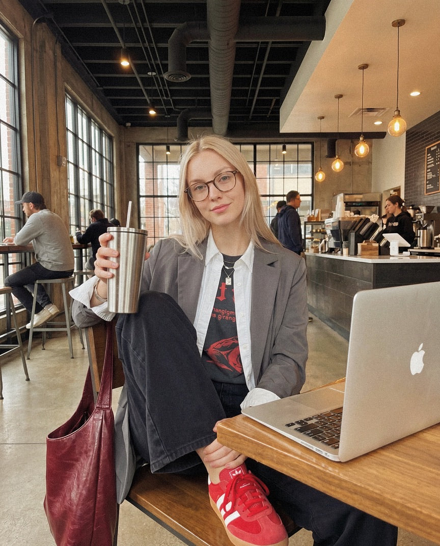 Student with glasses holding a tumbler at a cafe table with a laptop in front of her.