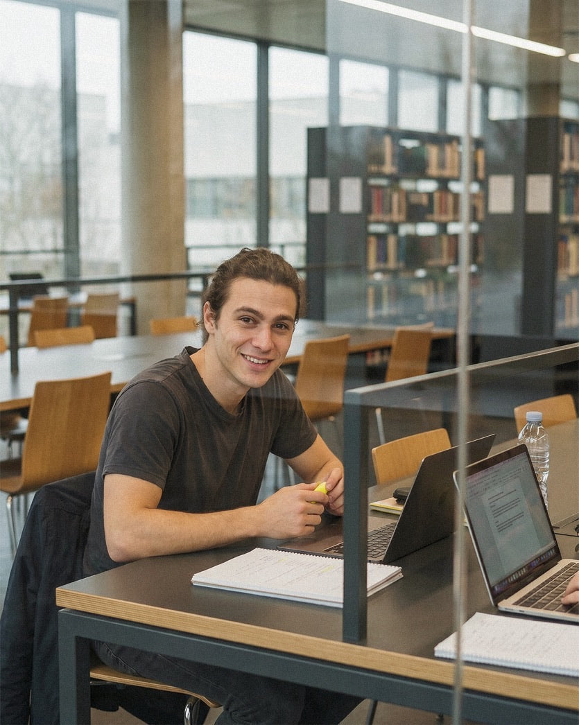 Student studying with laptop in a sunlit university library with bookshelves behind.