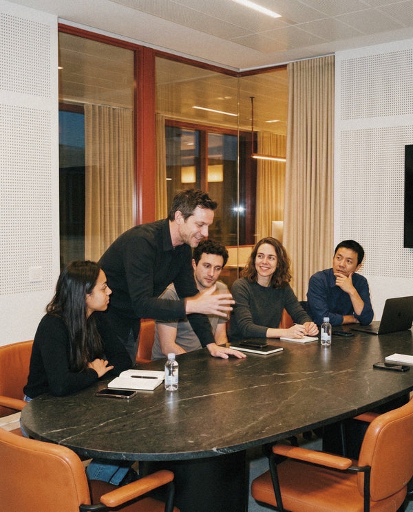 Five colleagues around a long meeting table with one person standing pointing, evening office light.