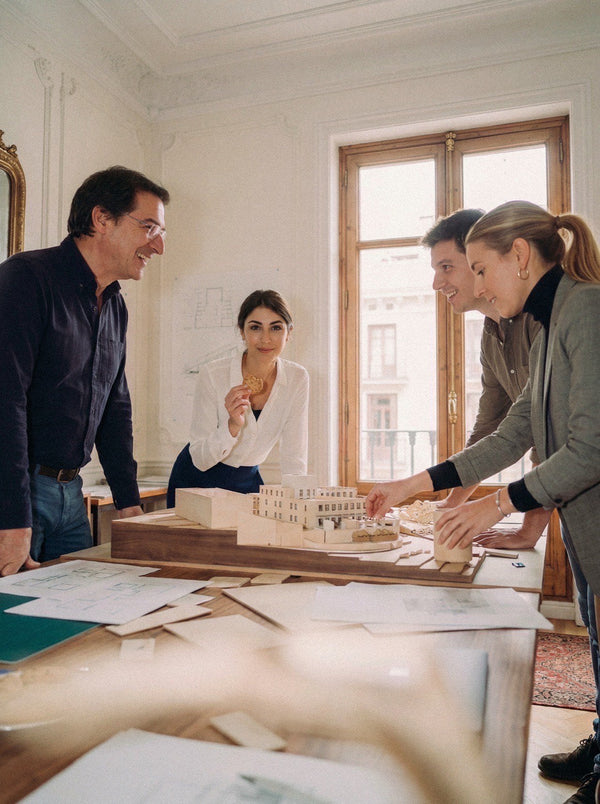 Four professionals examining an architectural model on a table in an ornate room with tall windows.