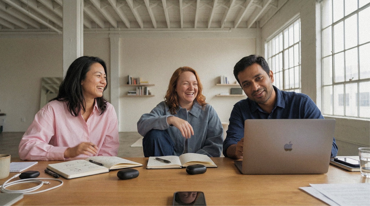 Three colleagues laughing around a workshop table with laptops, notebooks, and tall windows behind.