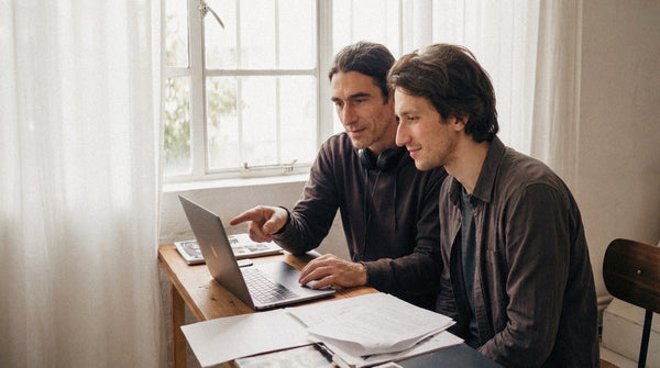 Two men collaborating at a laptop with one pointing at the screen and sketches on the desk.