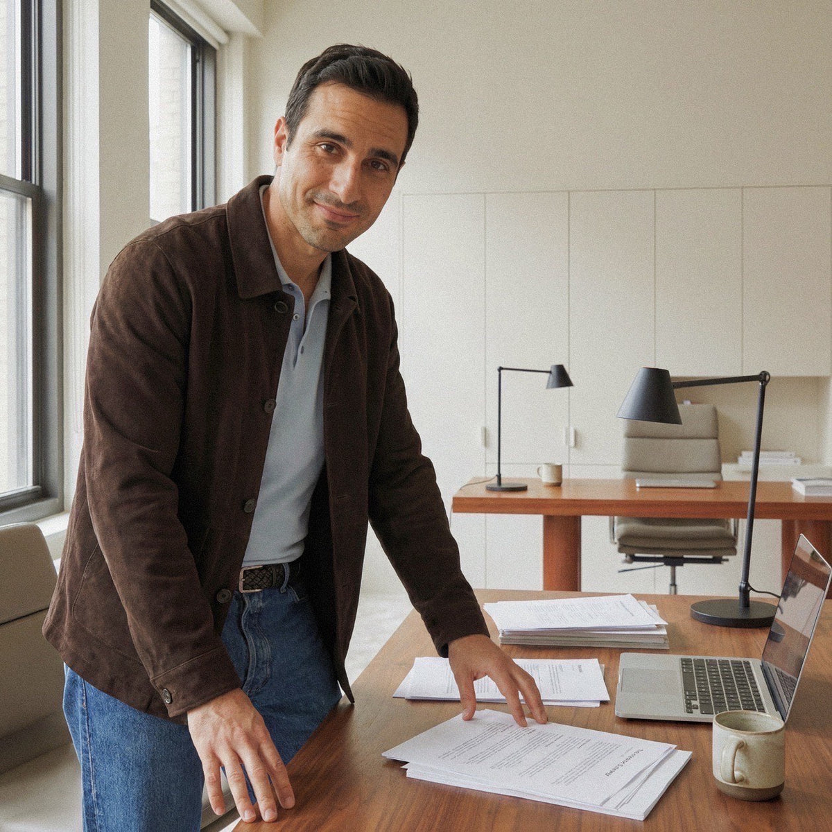 Professional in brown jacket leaning on a wooden desk with papers and laptop in a modern office.