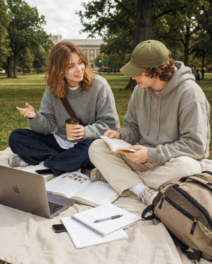 Two students studying on a campus lawn with laptop, books, and notes between them in front of a brick university building