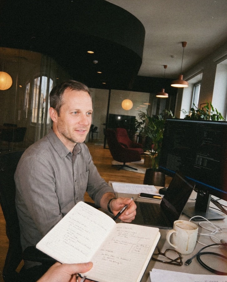 Man reviewing handwritten notes in an open notebook at a workspace with a monitor showing code in a warm-lit office