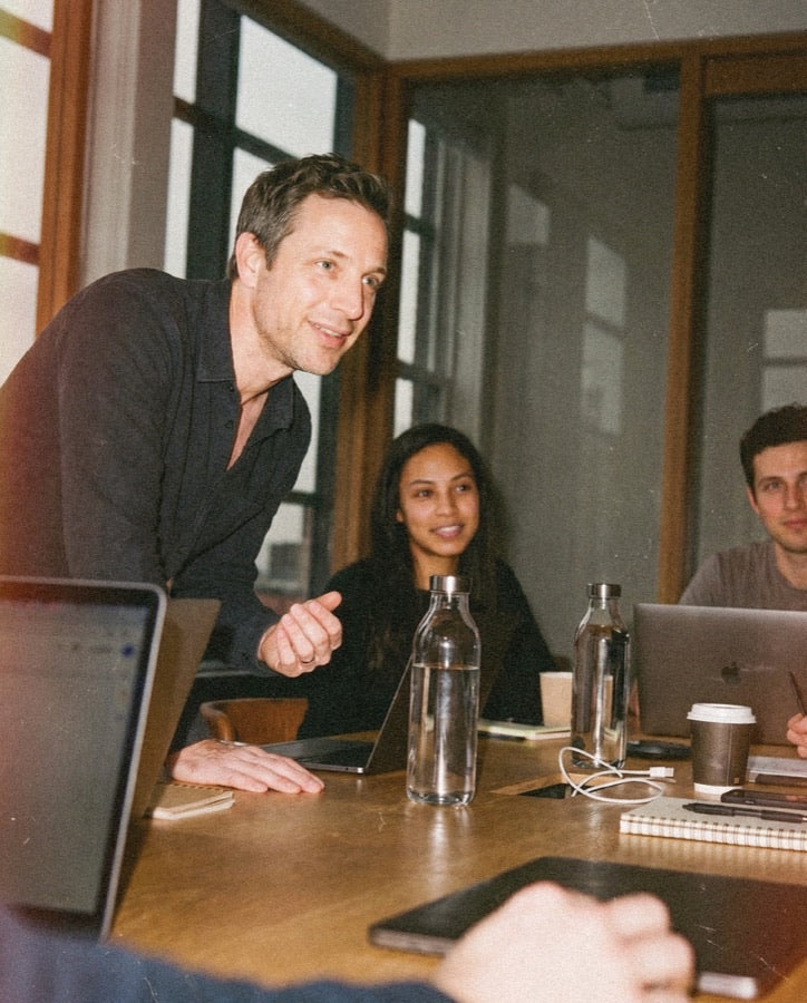 Man leaning forward at a wood meeting room table, gesturing to two colleagues with laptops and water bottles