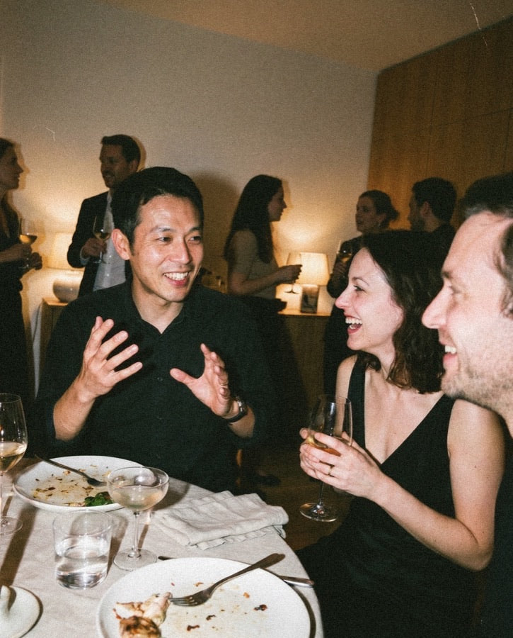 Three friends laughing together at a dinner party with wine glasses and shared plates on the table