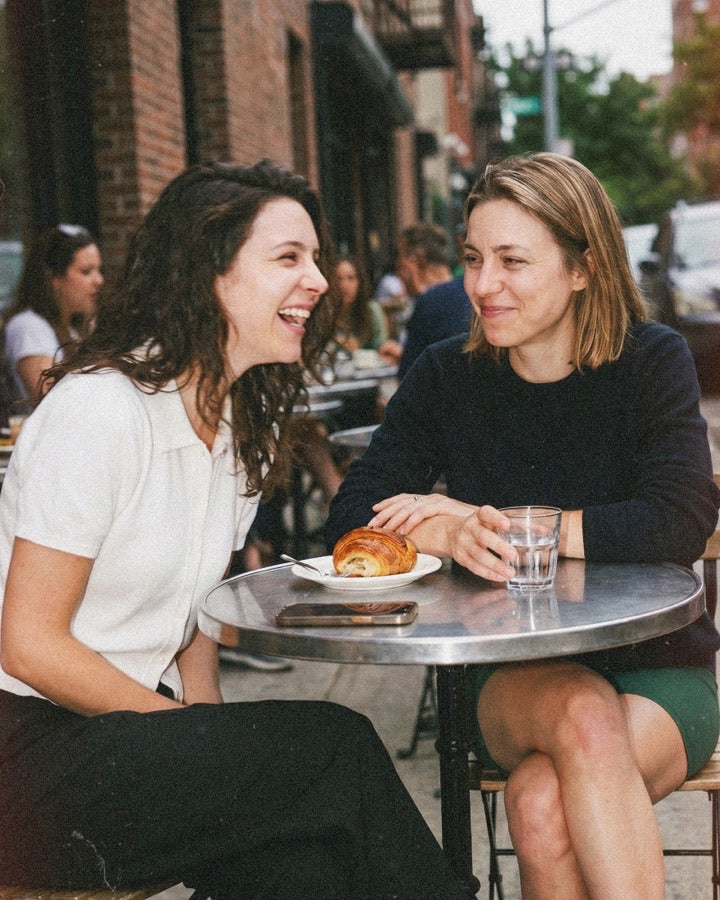 Two women laughing together at a sidewalk cafe table with a fresh pastry and a glass of water between them