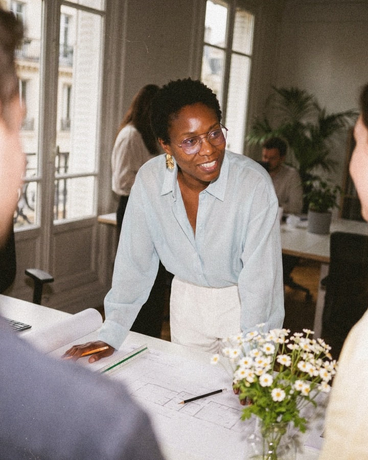 Woman in pale blue button-down shirt leaning over architectural drawings on a desk with daisies and colleagues in the background