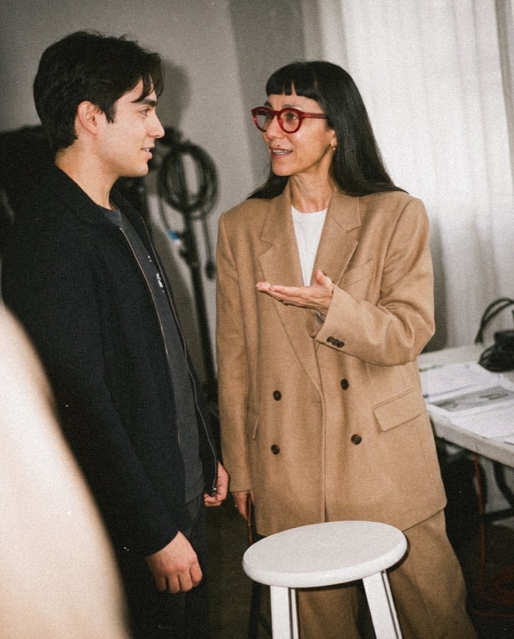 Woman in red glasses and tan blazer in conversation with a young man in dark hoodie, indoor studio with daylight