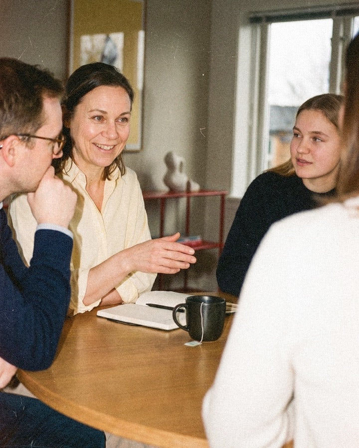 Four colleagues seated around a wood table in a warm room, woman in cream blouse speaking with a notebook open