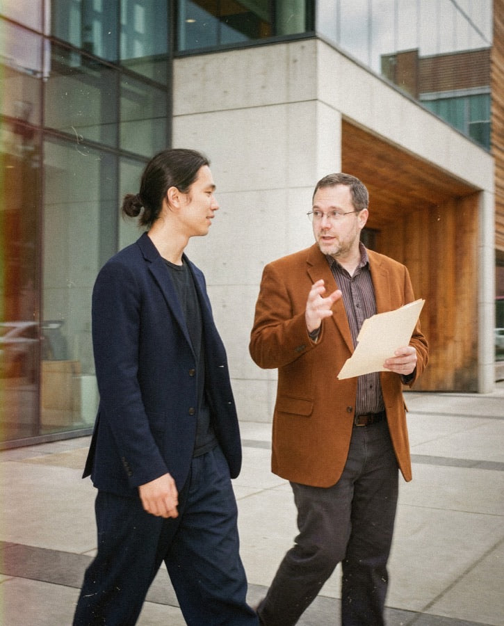Older man in rust blazer holding a folder, gesturing in conversation with a younger man outside a modern office building