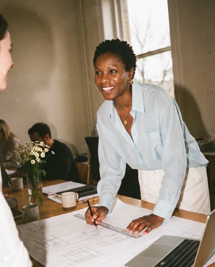 Person smiling and gesturing toward architectural plans on a sunlit wooden desk.