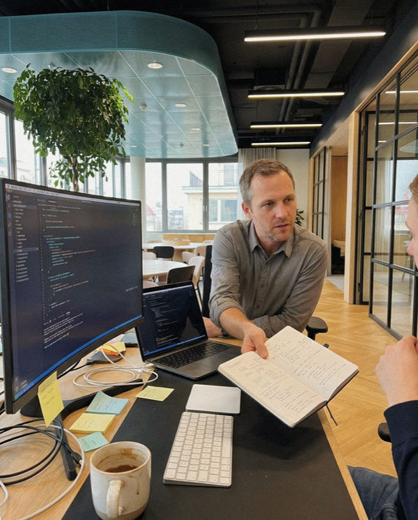 Man at a desk reviewing notes with a colleague, code on the monitor behind