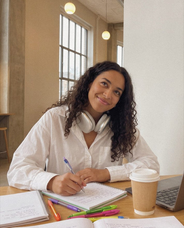 Smiling woman writing in a notebook with colorful pens at a sunlit table