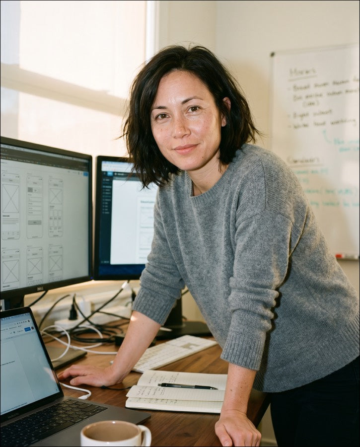 Woman at a desk with wireframe designs on dual monitors, looking at the camera